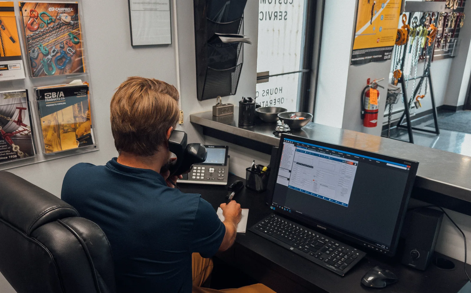 A Nelson Wire Rope salesman taking a call at his desk in the Hatfield office, notepad in hand and the order system open on the monitor beside him
