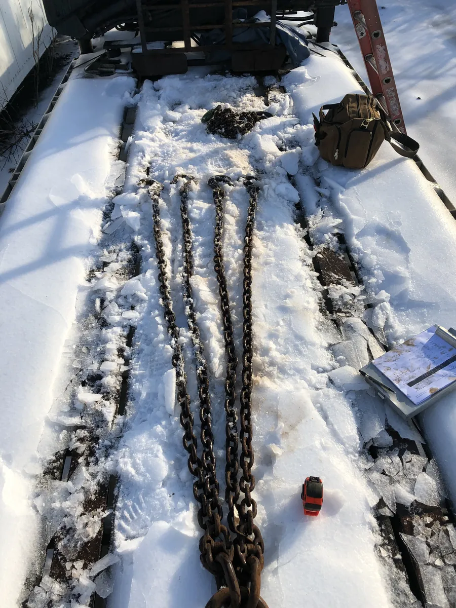 Chain slings laid out for inspection on a snow-covered flatbed trailer, with inspection paperwork alongside