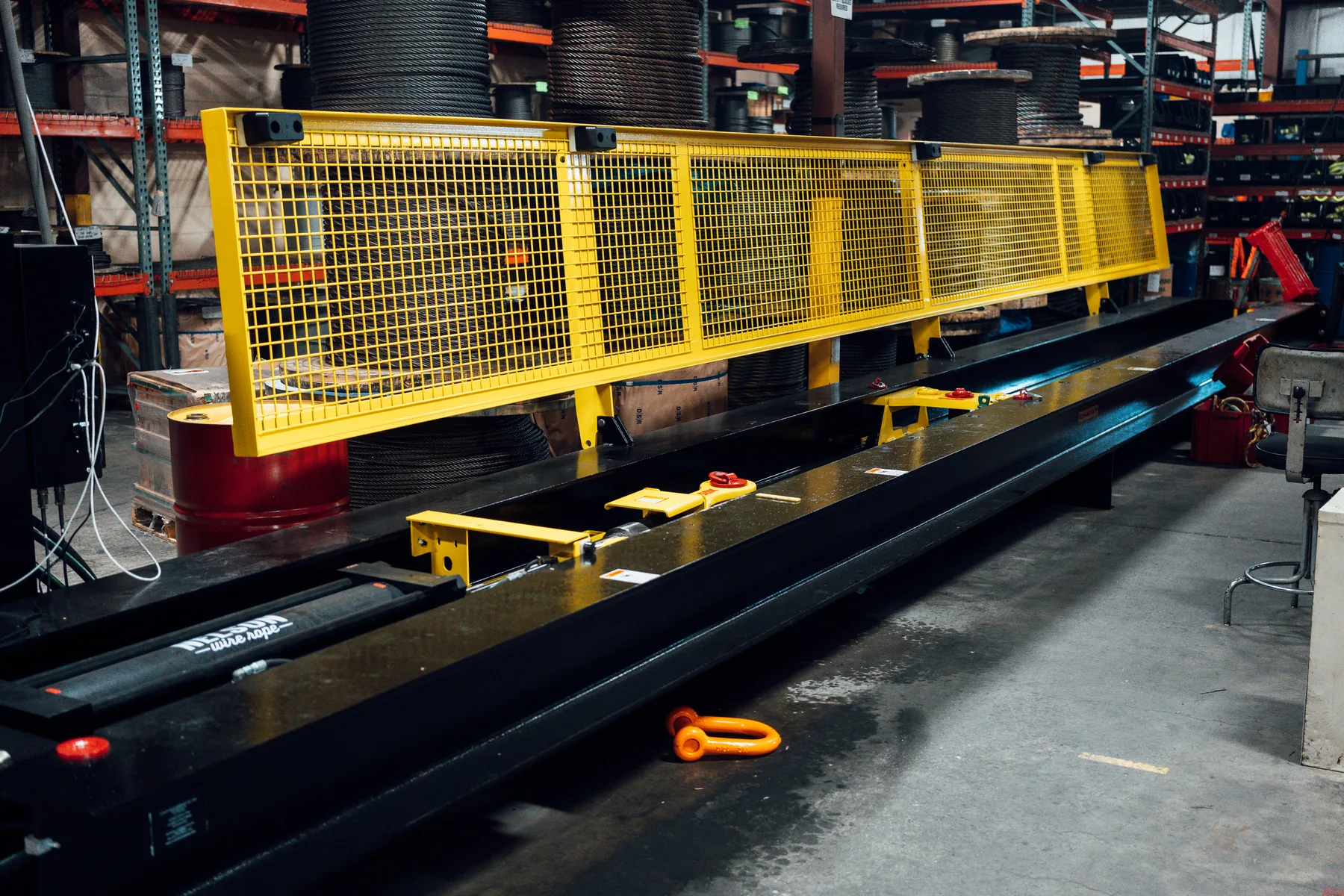 The horizontal proof test bed in the Nelson Wire Rope shop, a long steel bed with a yellow guard running its length, wire rope spools racked behind it