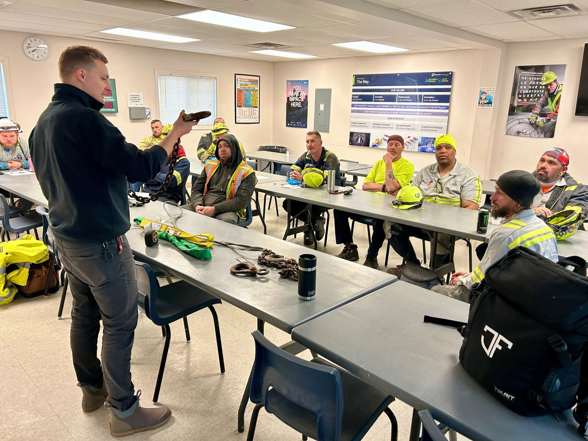 Steve Nelson holding up a worn chain sling hook in front of a seated crew, with a green round sling, a wire rope sling and a length of chain laid out on the table for the session